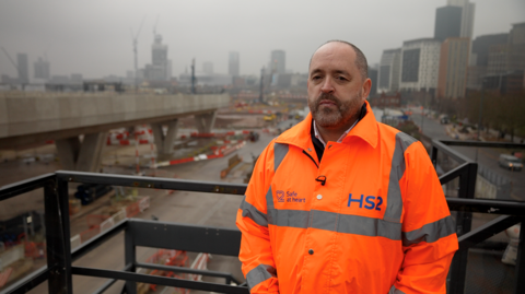 A man with very short dark hair and dark facial hair is wearing an orange high-vis reflective jacket that has the HS2 logo on it. He is standing on a platform that looks over a large viaduct and other construction work with high rise buildings in the background