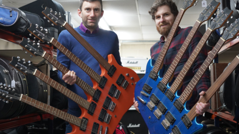 Two five neck guitars, one orange the other blue being held by two men looking at the camera with a Marshall amplifier in the background