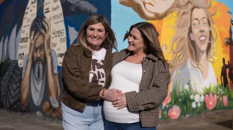 Wendy Robinson pictured with her daughter Amy standing in front of a colourful mural on two sides of a wall. Amy, on the right, has blonde hair and is smiling. There are blue skies, sun, flowers and birds behind her. On the mural towards the left is a picture of a man holding his head in his hands with words including 'cancer, stress, no, health' written in a lung-diagram on a dark blue background.