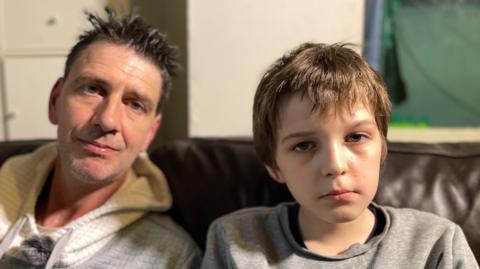 Dad with spiky hair and a white and grey hoodie with 12-year old son in a grey jumper on a brown leather sofa.