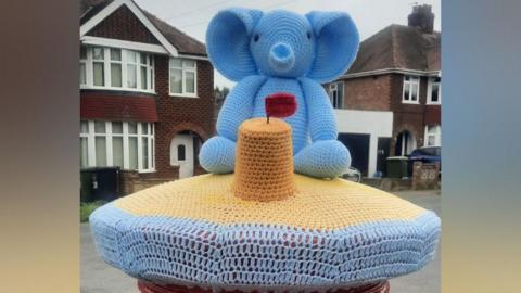 A knitted topper sits on top of a red post box. The topper features blue wool to represent the sea, followed by beige wool, depicting sand. On the sand, is a small knitted sandcastle with a red flag, and behind it is a blue knitted elephant.