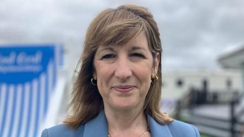 Rachel Reeves standing a smiling at the camera. She is wearing a gold necklace, gold hoop earrings and a light blue blazer.