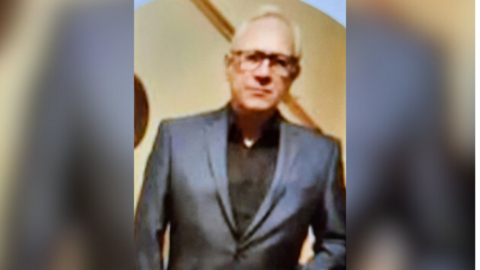 Blurry image of a man with white hair and wearing glasses and a grey suit.