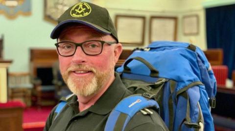 A photo of a man called Chris Jones. He is wearing a dark green hat with a yellow logo on it. He is also wearing dark rimmed glasses. He has a blue backpack on.