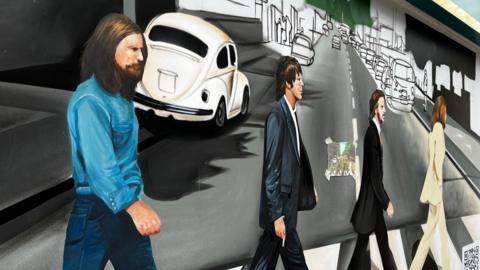 A mural depicting the four band members of The Beatles walking over a zebra crossing. One is in a white suit, two are in dark suits and the fourth is a denim top and jeans. Cars are depicted in the background. 