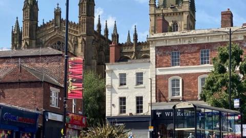 Wakefield city centre - with Wakefield Minster on the left, and shops, bushes and a bus stop on the right, and blue sky above