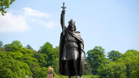 A bronze seen in front of a blue sky and green trees depicts King Alfred the Great holding a sword aloft with his right arm.