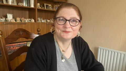Paula is looking at the camera and smiling. Her hair, which is auburn coloured, is pulled back into a pony tail behind her head. She has blue eyes. She has silver hooped earrings in. She is sat on a brown wooden chair. In the background is a brown wooden cabinet with cubby holes. There is lots of different little decor houses on the shelves.