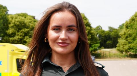 Nicole Douglas has long brown hair. She is standing in a car park where ambulances are parked on the left. She is wearing green NHS call handler uniform.
