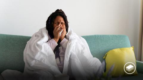 A photograph of a girl on a green sofa, with a blanket wrapped around her. She is sneezing into a tissue.