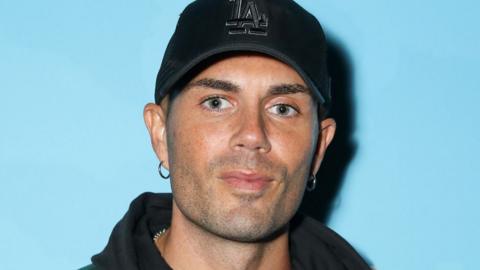 Max George wearing a black baseball cap standing against a light blue background in September 2023