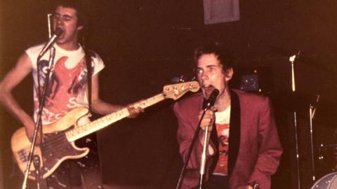 A vintage photo shows two musicians performing energetically on stage with microphones and instruments. One person plays a bass guitar while singing, and the other grips a microphone, leaning forward mid-performance. The dim lighting and grainy texture give the image a raw, punk-rock concert atmosphere.