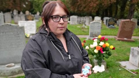 Jasmine Beverley wearing a black coat and black glasses holding her premature baby's small blue teddy bear. She is stood in front of her son's grave which is full of coloured roses.