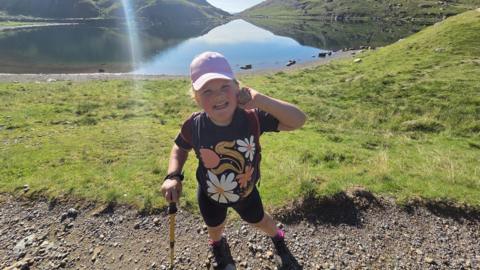 A six-year-old girl wearing a navy blue t-shirt, shorts and a pink baseball cap. She is stood on a footpath with an area of grass and a lake behind her.