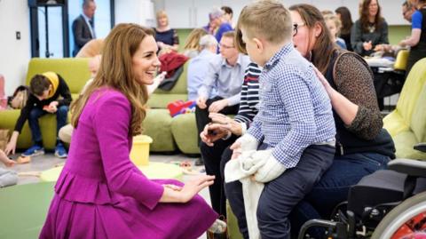 Princess of Wales wears a magenta skirt suit and is kneeling while chatting to a child and his carer. Other adults and children are in the room.