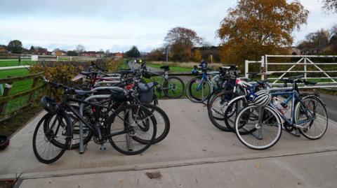 A number of bicycles are locked to two lines of bicycle racks on a small patch of concrete. To the right side, a small road leads to a white metal gate, and there is wooden fence behind and alongside the bikes. Behind that is a green racecourse with some trees, and houses in the background.