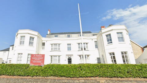 A white building with multiple windows. A brick wall and hedge are in front of it. There is a red sign with reads "St Aubyn's"