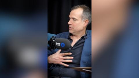 A man holds his hand over his chest. He is wearing a dark shirt with a blue jacket over it.