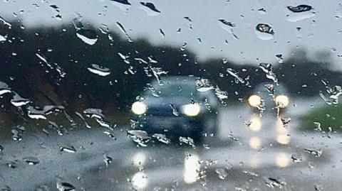 A photograph taken through a window covered in raindrops. Two cars driving with headlights on are visible. 