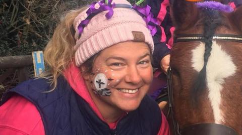 Hazel Ackland smiles at the camera as she crouches next to a brown coloured pony. She wears a pink beanie hat with some purple decorations attached to it. She also has some face paint on her cheeks. 