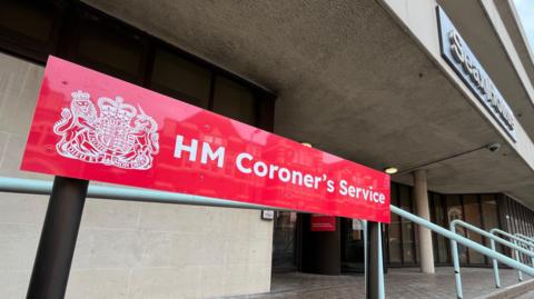A red sign that says HM Coroner's Service. It is at the entrance to Essex Coroner's Court, which is a cream building that has a black revolving door and brick paving outside.