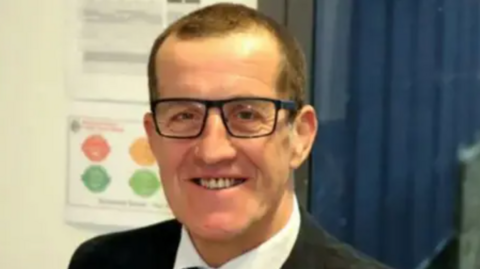 A man with short cropped dark hair and wearing a suit and glasses smiles at the camera