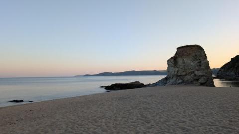Carlyon Beach at sunset is empty of people but the sand shows many footprints. The sky is pale shades of pink, orange, yellow and blue. There is a large boulder on the shoreline and beyond the sea a headland is in silhouette.