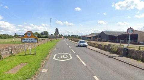 Google Maps image of the B6412 leading into Temple Sowerby. A sign on the right states the name of village as well as a 30mph speed limit. A farm building stands on the right, as does a light-coloured saloon car which is parked half on the pavement. The sky is blue with a line of fluffy white clouds and tall trees in the distance are covered in leaves. To the left, behind the road sign, a large group of sheep can be seen in a grassy field.