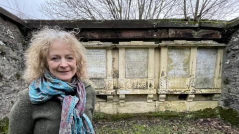 Jan Hogarth, who chairs the Friends of Wamphray Church steering group,  stands in the Rogerson family crypt. She has shoulder length, blonde, wavy hair and is wearing a dark green jumper and a large blue and purple silk scarf. 