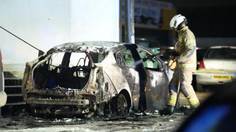 A car is scorched as a firefighter sprays it with water. 