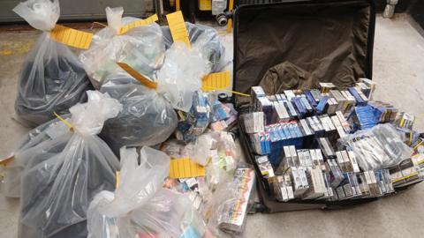 Full plastic bags and a suitcase full of cigarette cartons and vapes on the ground. The bags have been labelled with yellow cards.