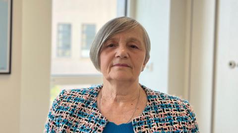 Mary has short grey hair wearing a patterned jacket over a blue top, photographed indoors in an office setting. A framed map artwork hangs on the wall behind, and a window lets in natural light.