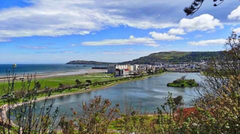 An ariel view of Mooragh Park on a sunny day with largely blue skies. There is a large lake with a small island in the middle and there are houses in the background in front of a grass and tree-covered hill.