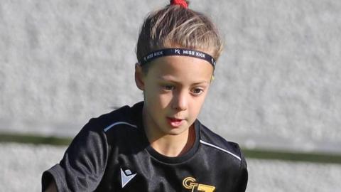 Poppy Atkinson is pictured standing in front of a stone wall. She is wearing a black sports top and has her hair tied up. There is a look of intent on her face.