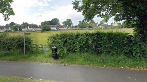 A public footpath sign pointing across a grassy field next to a metal gate and a black litter bin next to it. Houses are in the distance. 