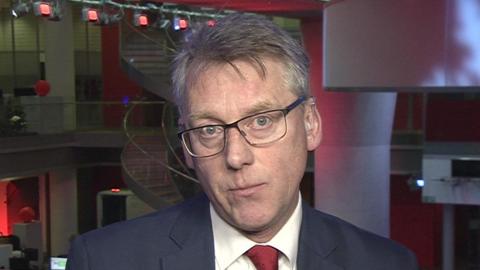 Sir Julian Hartley looking into the camera. He wears glasses, a blue suit jacket, white shirt and red tie