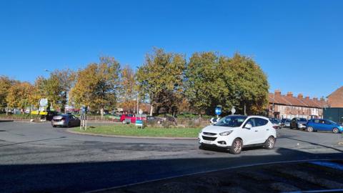 The Cross Rifles roundabout in Bridgwater. There are cars driving around it. Trees and buildings can be seen in the distance.