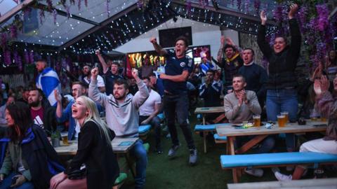 Football fans watching a game while in a pub's beer garden, with drinks on tables in front of them.