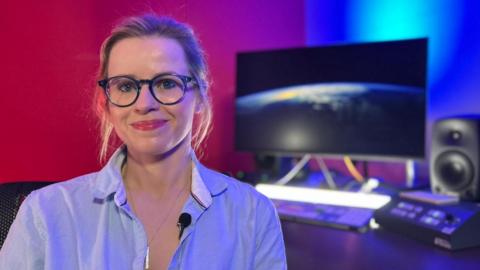 Christina Mackaill, who is wearing glasses and a blue shirt, smiling at the camera. There is a computer screen with an image of the moon in the background.