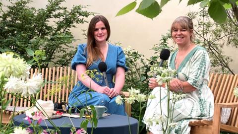 Two woman sat in a leafy garden, smiling at the camera. 