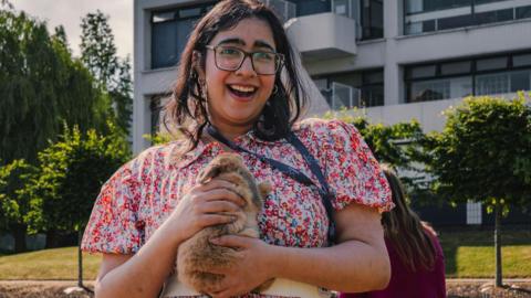 A young woman pictured outside holding a brown rabbit with two hands close to her chest. She is wearing a pink floral dress. She has shoulder-length dark hair and is smiling. 