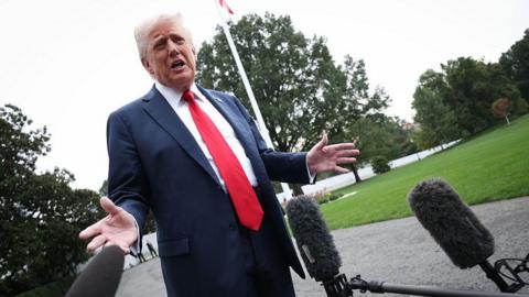 Trump speaking into microphones outside White House, he's wearing a suit with red tie.