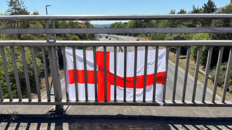 A St George's flag pinned to the railings on a bridge above a dual carriageway. The flag is on the outside of the bridge with the photo taking from behind it on the bridge. It is a sunny day with a clear blue sky and many trees lining the road.