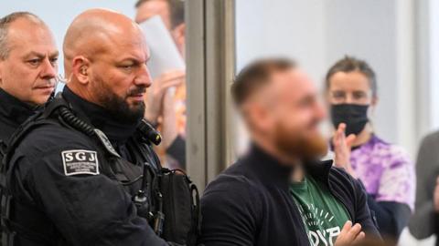 Defendant Johann G (blurred and on the right)  is brought to the courtroom at the start of a trial against seven left extremists accused for membership in a criminal organization on November 25, 2025 at the Higher Regional Court of Dresden, eastern Germany