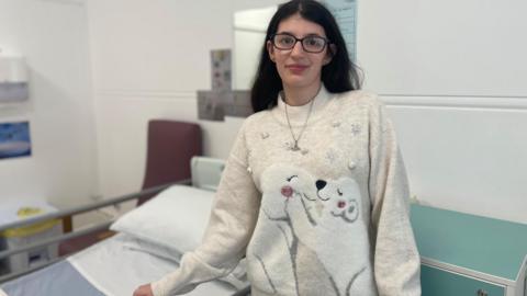 A young girl in a white jumper stands infront of a hospital bed