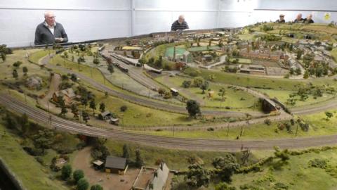 The model railway has green fields, tracks and buildings. There is a white wall in the background.