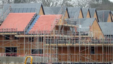 Houses being built. The homes have a both grey and red slate roofs and are made of orange bricks. In front there is scaffolding.