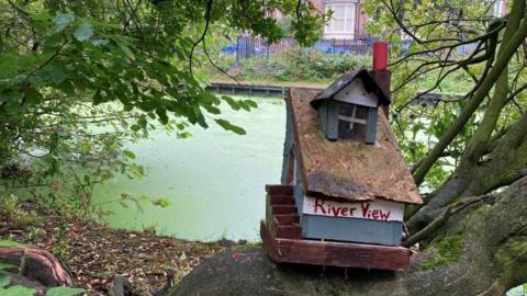 A small model house sits on a tree beside a river, with 'river view' painted in red on the white front wall. 