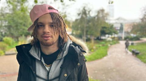 Lorenzo Alara looks into the camera in a park, which is blurred in the background. He wears a pink baseball cap backwards on his head, as well as a coat over a grey zip-up hoodie with a gold chain around his neck