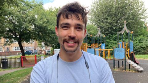 Matt Hollinshead, pictures smiling in front of a playground. He has dark hair and is wearing a light-coloured t-shirt and a pair of headphones around his neck. 